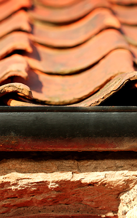 A close up of the edge of a roof showing a cast iron rhone with bubbling black paint and red pantiles rising in ridges behind it.