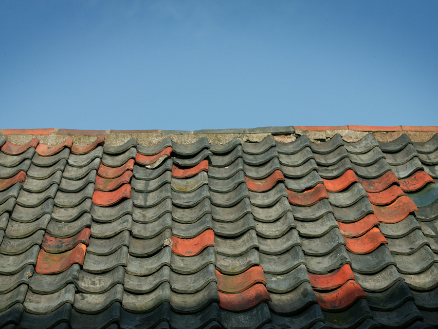 A close up shot of a roof with mostly dull black pantiles, lifted with the odd red one.