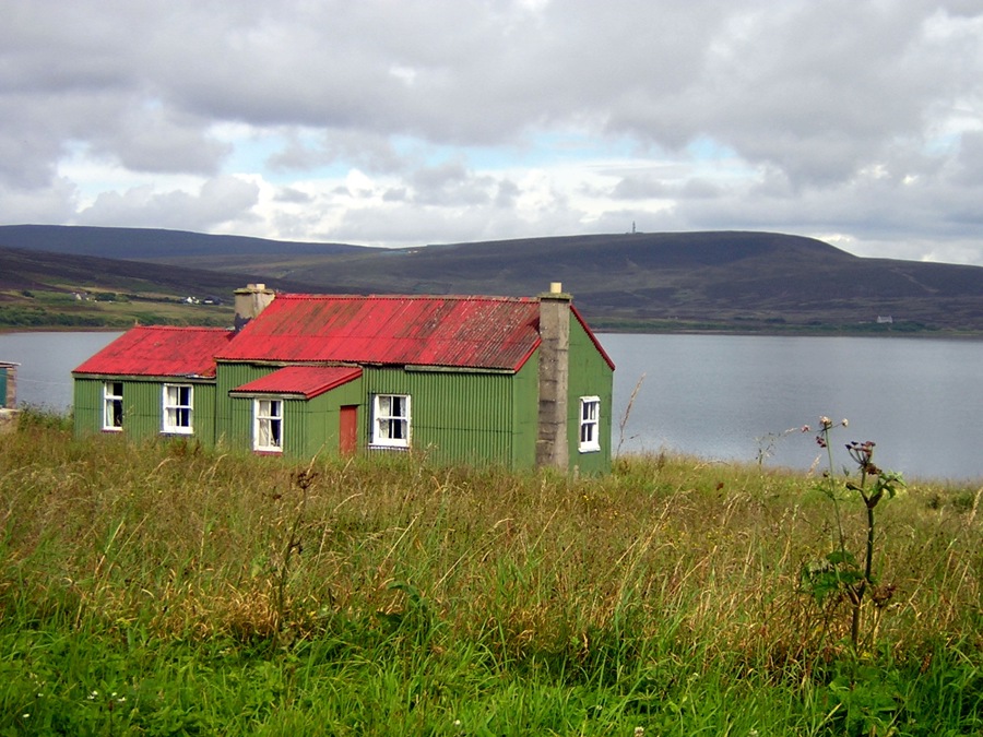 A green corrugated iron house with an orange corrugated iron roof at the edge of a loch