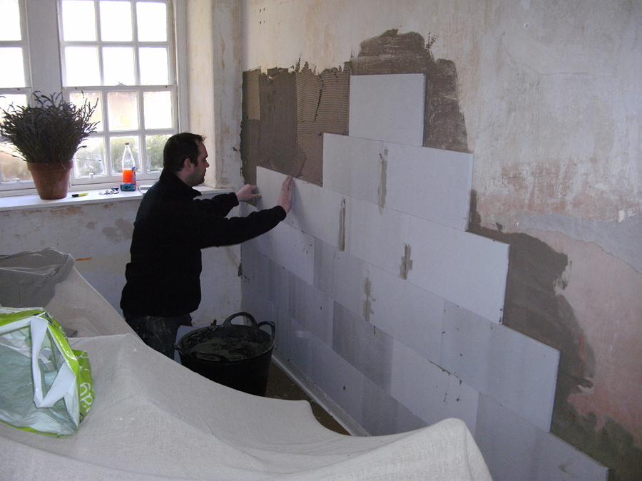 A person dressed in black clothing applies light-coloured rectangular insulation tiles to an interior wall.