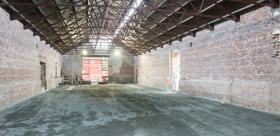 A large empty building with a concrete floor, exposed brick walls and large, exposed ironwork roof