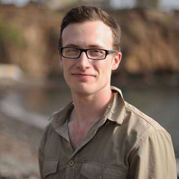 A person standing outside, with square glasses and short brown hair, wearing a brown open neck shirt, smiling