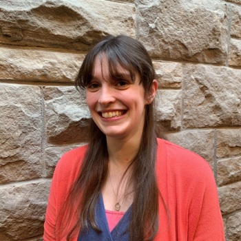 A headshot of Joanna smiling, standing against a brick wall, wearing a red cardigan