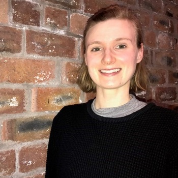 A headshot of Monica smiling, standing against a brick wall