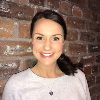A headshot of Rachel smiling, standing against a brick wall