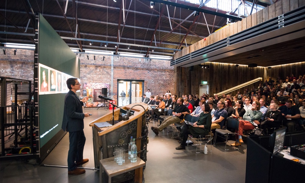 A speaker standing in front of a lectern and giant screen inside the Engine Shed