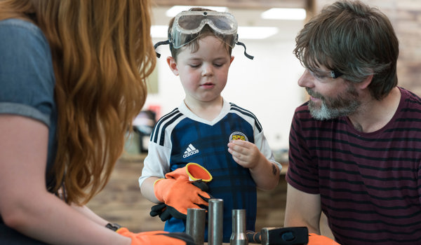 A child wearing safety glasses looking at a metal coin they made with two adults