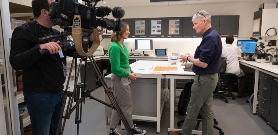A camera man holds a large broadcast film camera on a tripod. In front of the camera, there are a number of professionals talking and working in a lab.