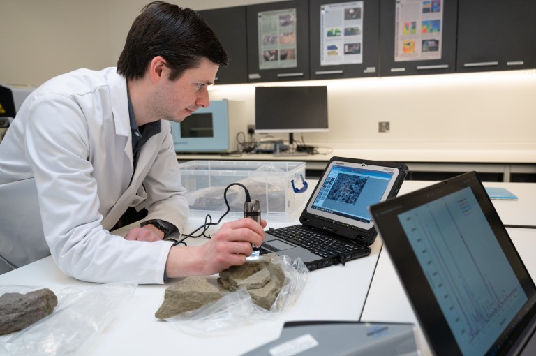 A heritage scientist in a white coat in a lab, scanning a piece of stone in front of a laptop