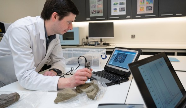Staff member in a white lab coat, standing in a lab. Looking at a laptop and pieces of stone. 