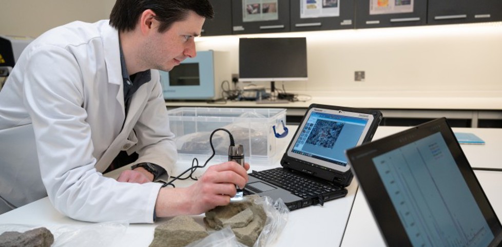 Staff member in a white lab coat, standing in a lab. Looking at a laptop and pieces of stone. 