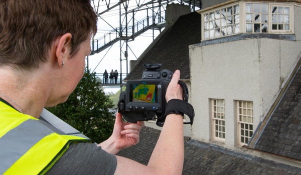 A scientist in high vis is looking at the roof of the Hill House using a thermal camera. The picture on the camera screen is showing a thermal image of yellows, reds and blues.