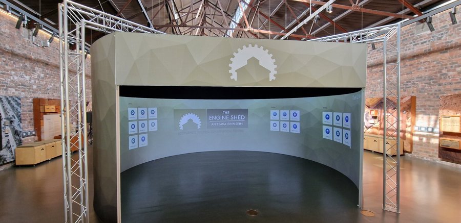 A semi-circular, free-standing hub inside the Engine Shed, displaying the Engine Shed logo on a wrap-around circular screen