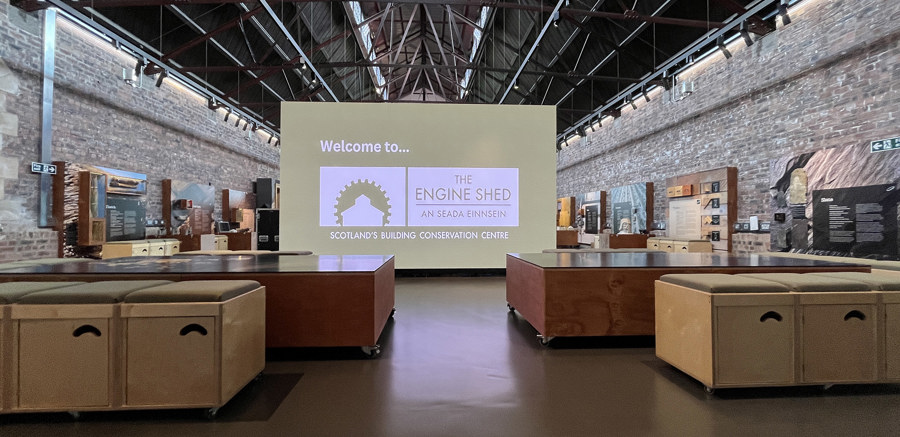 An interior view of the Engine Shed building. Shows an exhibition space, small wooden box chairs and a large LED screen which says Welcome to the Engine Shed.