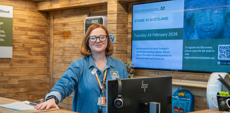 A member of staff at the Engine Shed is standing behind a reception desk, smiling towards the camera. The desk and reception surround is covered in rustic wooden panels. 
