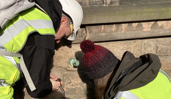 One person is holding a piece of equipment up to a stone building and the other is looking at the monitoring screen attached to the device.