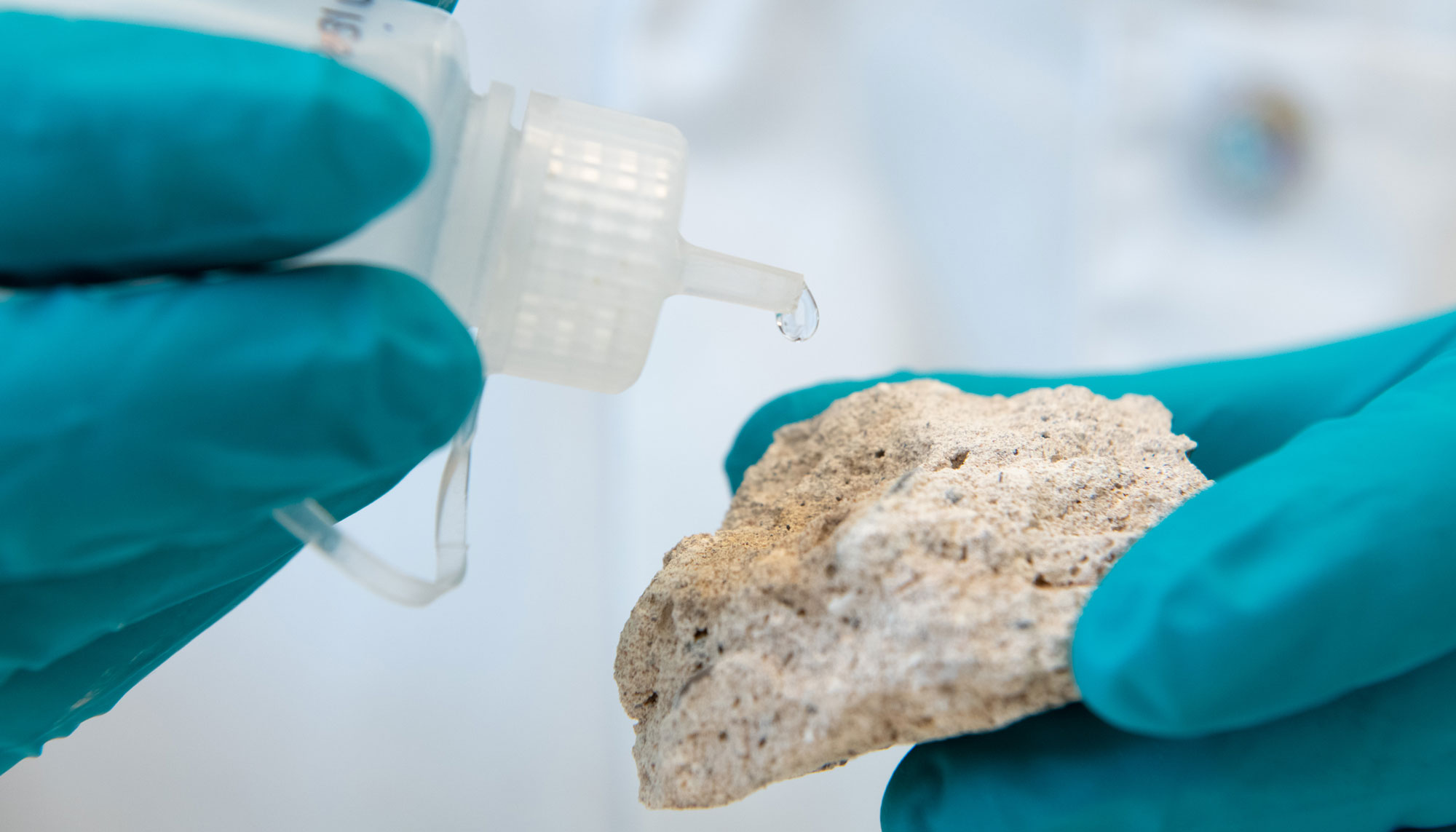 Two hands wearing PPE gloves, holding a mortar sample in one hand and plastic dropper bottle in the second.