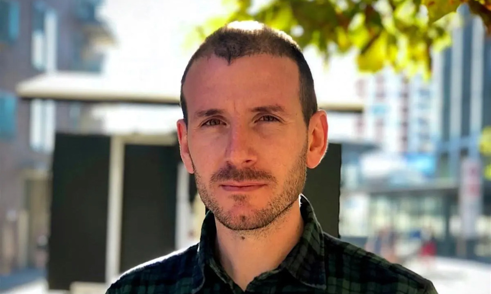 A person with a closely shaved head and moustache, wearing a checked black and green shirt standing outside a building with a tree behind them