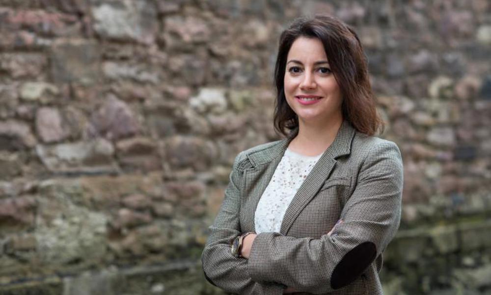 A portrait of Lila Angelaka in a formal pose, standing in front of masonry wall.