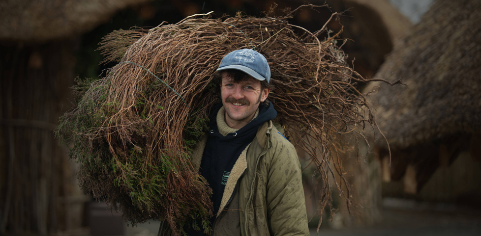  A craft fellow is standing smiling to camera. They are wearing a blue cap and green jacket. They are carrying a bundle of thatching materials on their shoulder. 