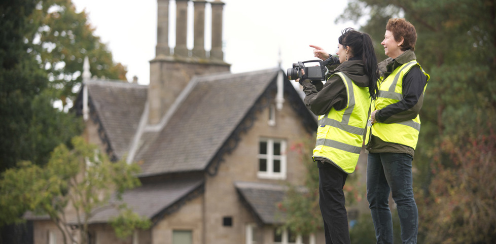 Two HES staff members wearing high-visibility yellow vests. They are standing in front of a traditional building, with equipment to survey it.