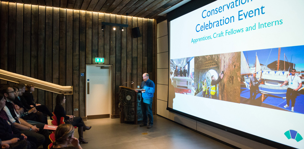 A view in the auditorium of the Engine Shed. A staff member stands in front of a large presentation screen and is giving a talk to an audience seated in auditorium.