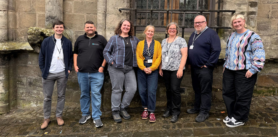 A group of 7 people standing in front of a traditional stone building. They are all wearing smart/casual clothing and smiling to camera. 