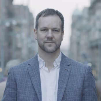 A person wearing a blue blazer and white shirt, with short brown hair and a short beard