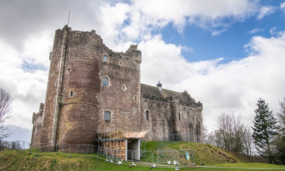 A large grey stone castle surrounded by temporary fencing with the entrance covered in scaffolding.